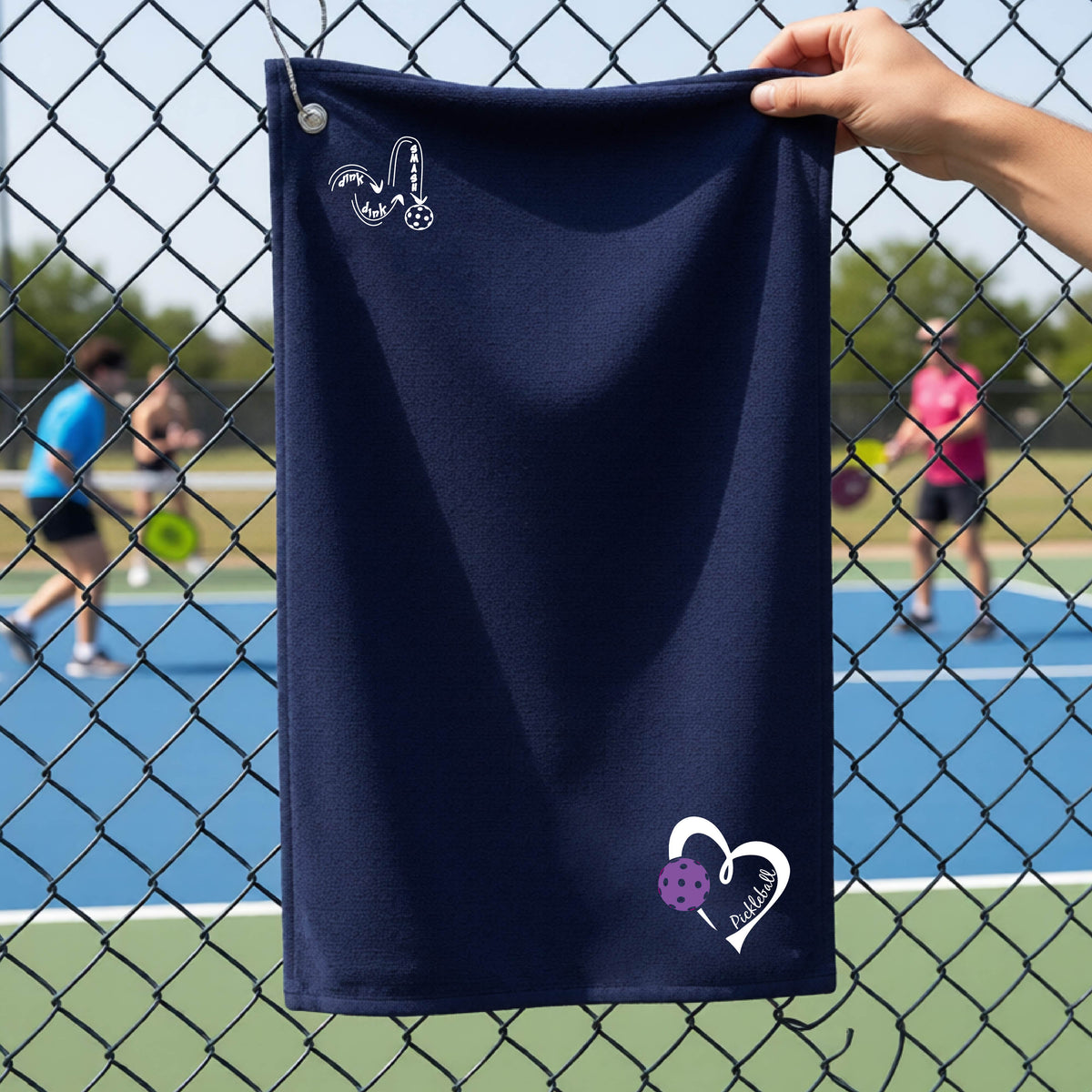 A navy blue terry cloth towel held up by a hand in front of a chain-link fence and outdoor pickleball court. The towel has a white stylized logo near the top left 'DINK DINK SMASH' and a white heart logo with a purple pickleball graphic and the word 'Pickleball' near the bottom right.