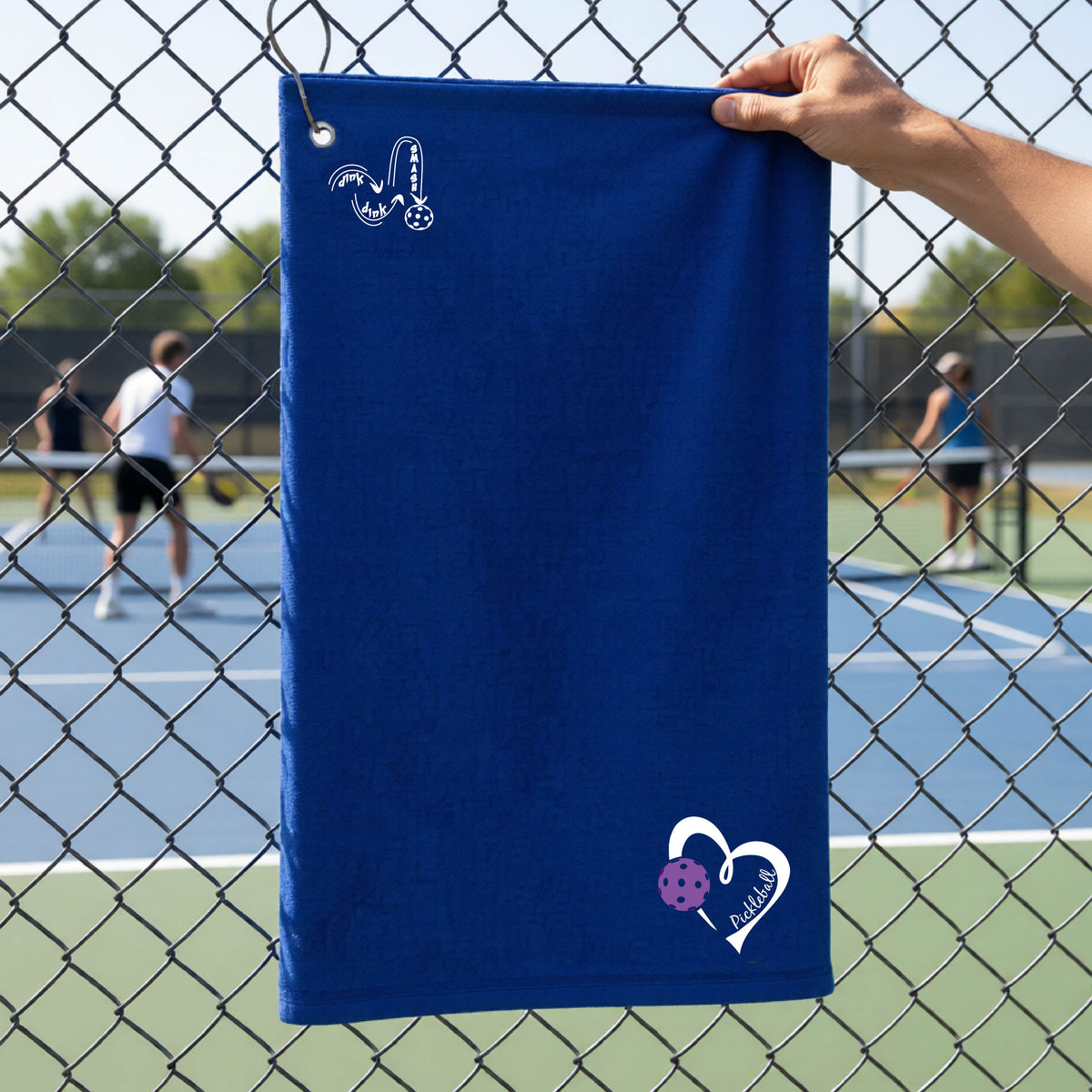 A royal blue terry cloth towel held up by a hand in front of a chain-link fence and outdoor pickleball court. The towel has a white stylized logo near the top left 'DINK DINK SMASH' and a white heart logo with a purple pickleball graphic and the word 'Pickleball' near the bottom right.