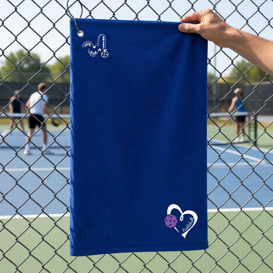 A royal blue terry cloth towel held up by a hand in front of a chain-link fence and outdoor pickleball court. The towel has a white stylized logo near the top left 'DINK DINK SMASH' and a white heart logo with a purple pickleball graphic and the word 'Pickleball' near the bottom right.