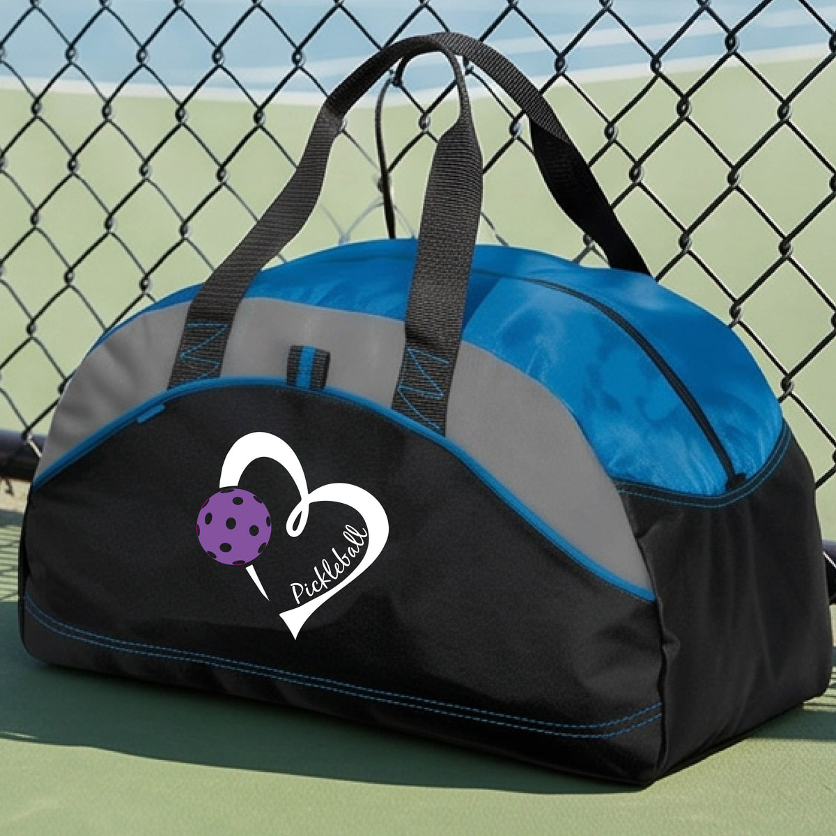 A black and royal blue pickleball duffel bag with a white heart logo and purple pickleball graphic, resting on a green pickleball court.