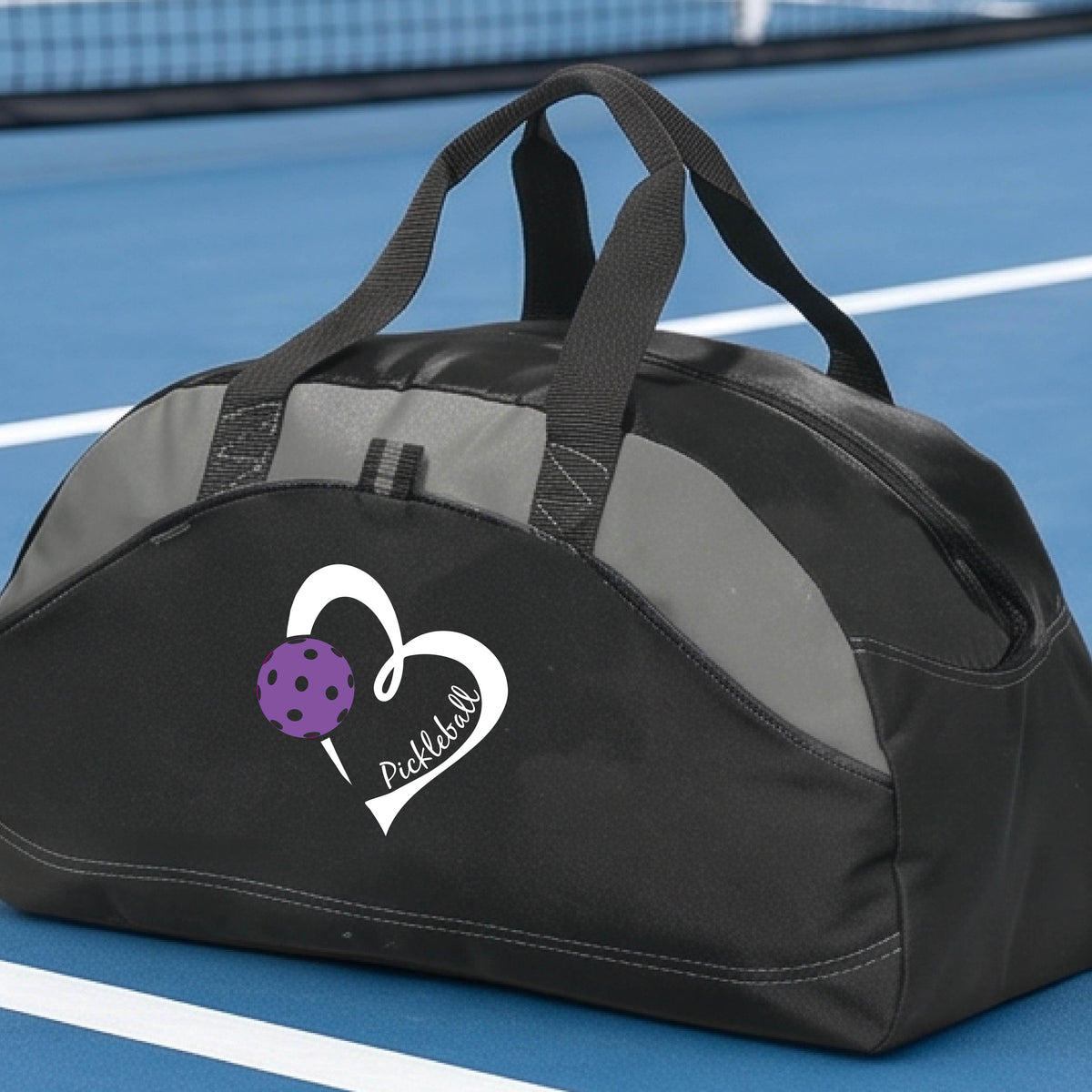A black pickleball duffel bag with a white heart logo and purple pickleball graphic, resting on a green pickleball court.