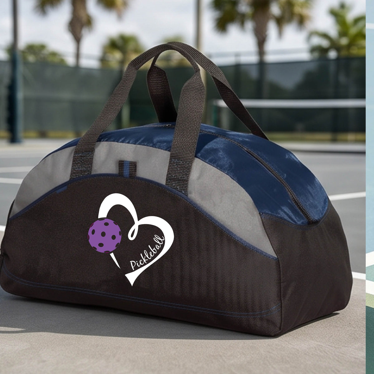 A black and navy blue pickleball duffel bag with a white heart logo and purple pickleball graphic, resting on a green pickleball court.