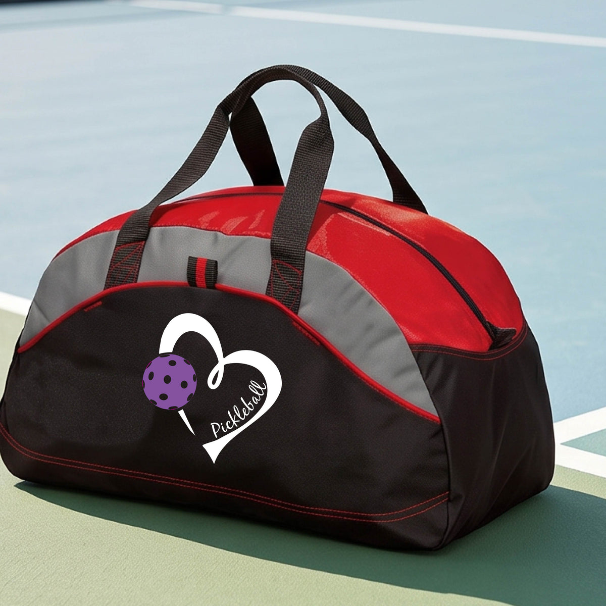 A black and red pickleball duffel bag with a white heart logo and purple pickleball graphic, resting on a green pickleball court.