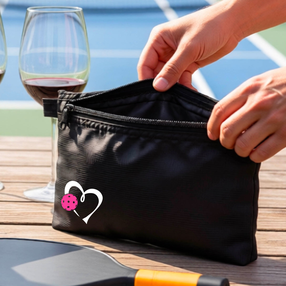 A black zippered pouch with a white heart and pink pickleball graphic, shown on a wooden table with a wine glass, paddle, and hands reaching into the pocket, with a pickleball court in the background.