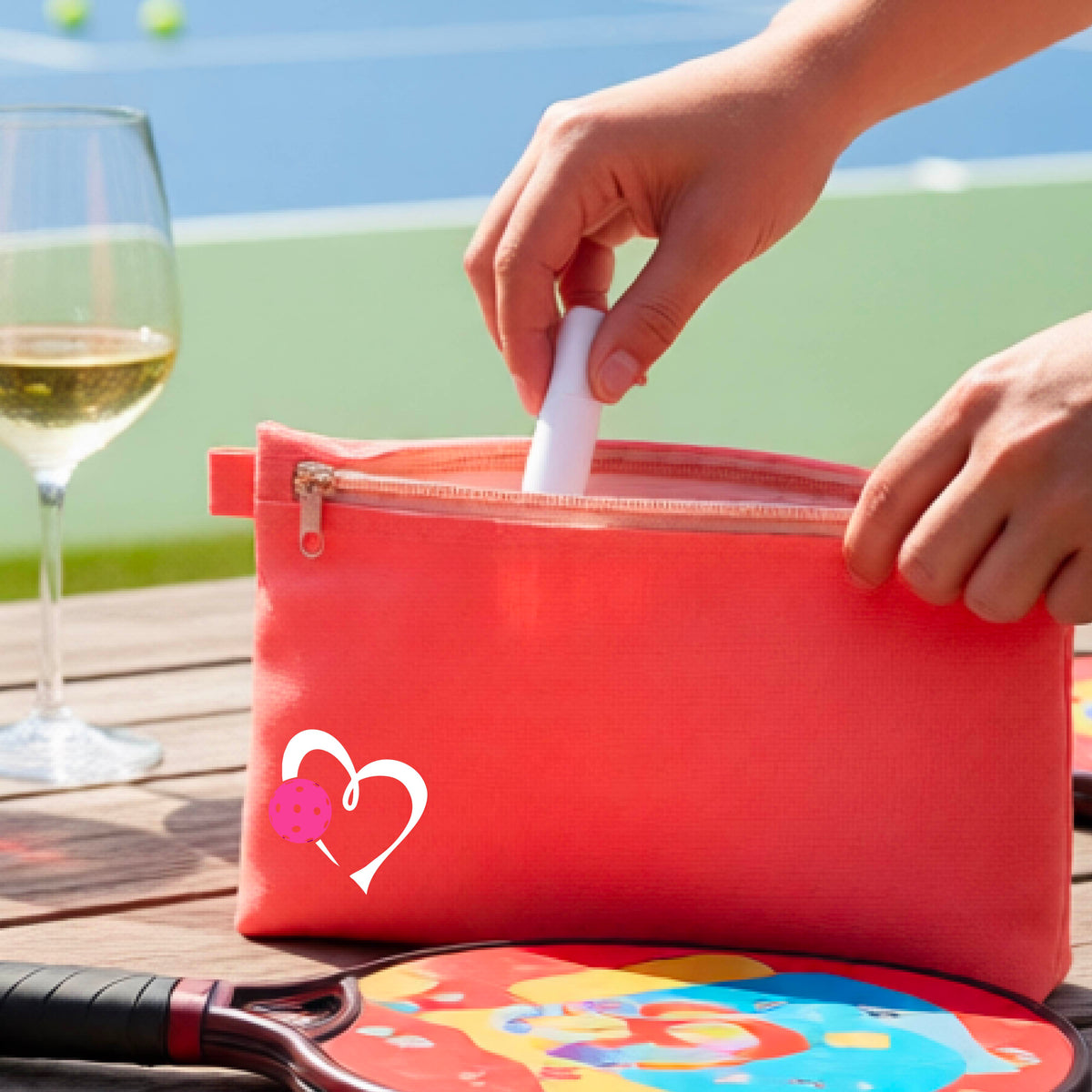 A coral pink zippered pouch with a white heart and pink pickleball graphic, shown on a wooden table with a wine glass, paddle, and hands reaching into the pocket, with a pickleball court in the background.