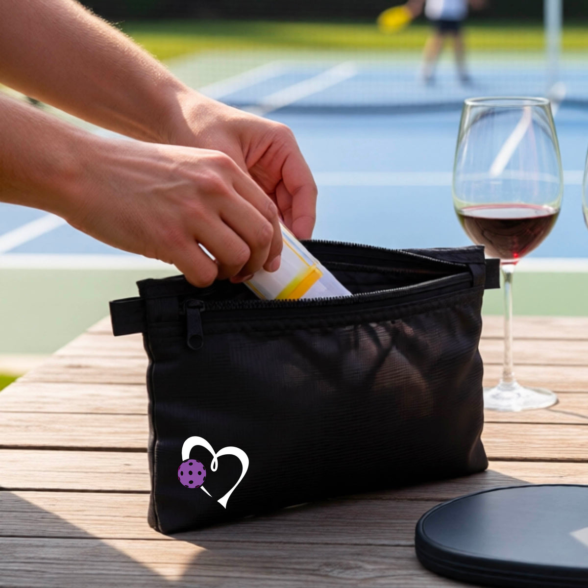 A coral pink zippered pouch with a white heart and purple pickleball graphic, shown on a wooden table with a wine glass, paddle, and hands reaching into the pocket, with a pickleball court in the background.