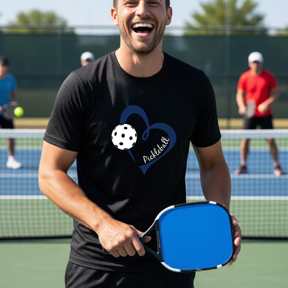 A man laughing on a pickleball court wearing a black t-shirt with a navy blue heart, white pickleball graphic, and the text 'pickleball' printed on the front.