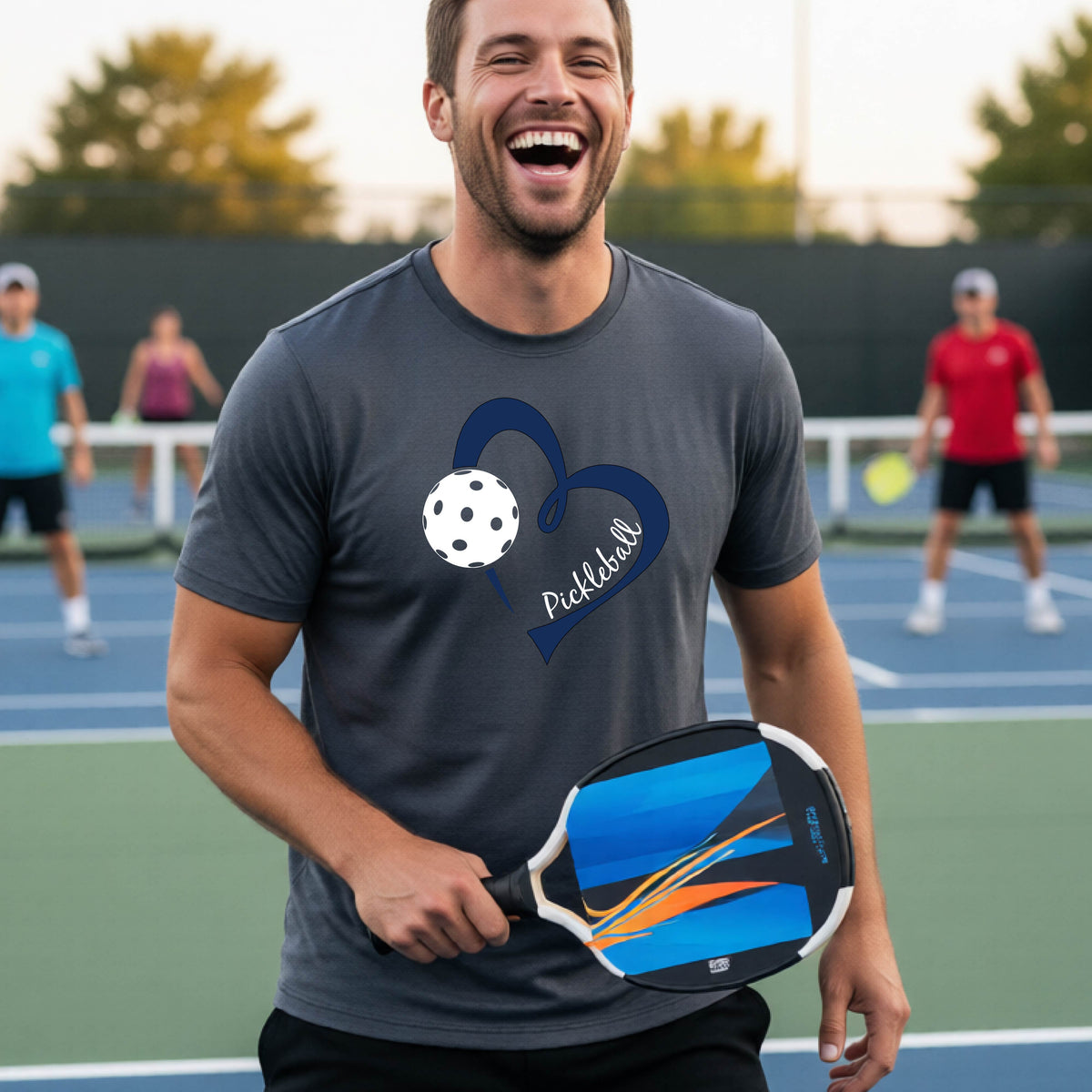 A man laughing on a pickleball court wearing a dark gray t-shirt with a navy blue heart, white pickleball graphic, and the text 'pickleball' printed on the front.
