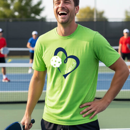 A man laughing on a pickleball court wearing a bright lime green t-shirt with a navy blue heart, white pickleball graphic, and the text 'pickleball' printed on the front.