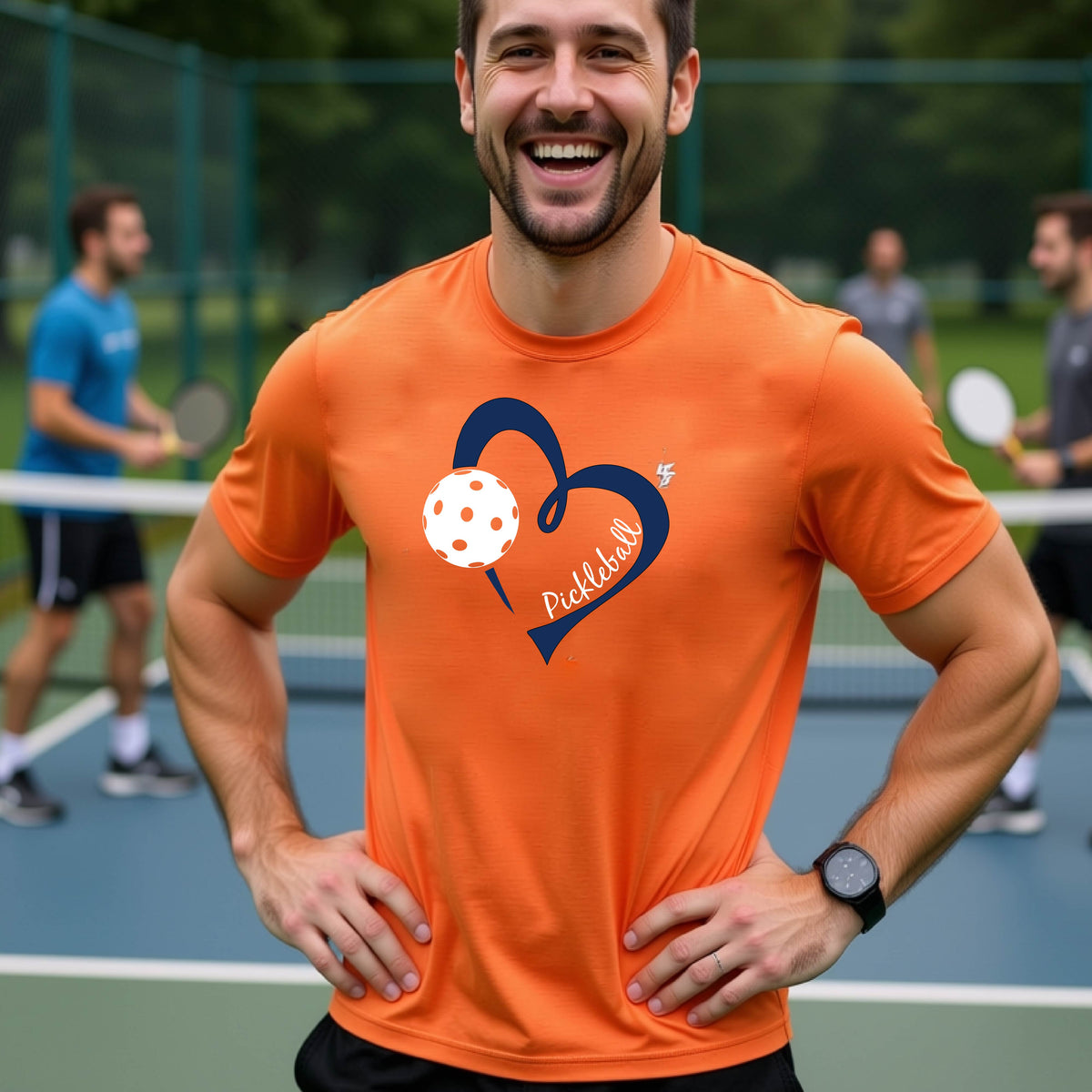 A man laughing on a pickleball court wearing a bright orange t-shirt with a navy blue heart, white pickleball graphic, and the text 'pickleball' printed on the front.