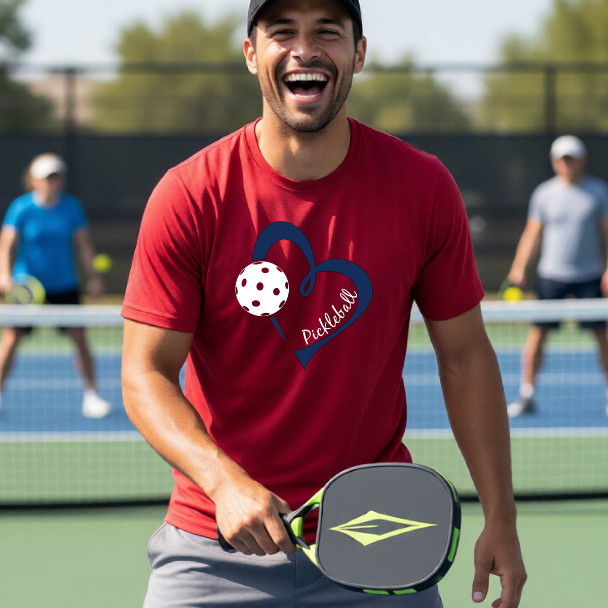 A man laughing on a pickleball court wearing a red t-shirt with a navy blue heart, white pickleball graphic, and the text 'pickleball' printed on the front.