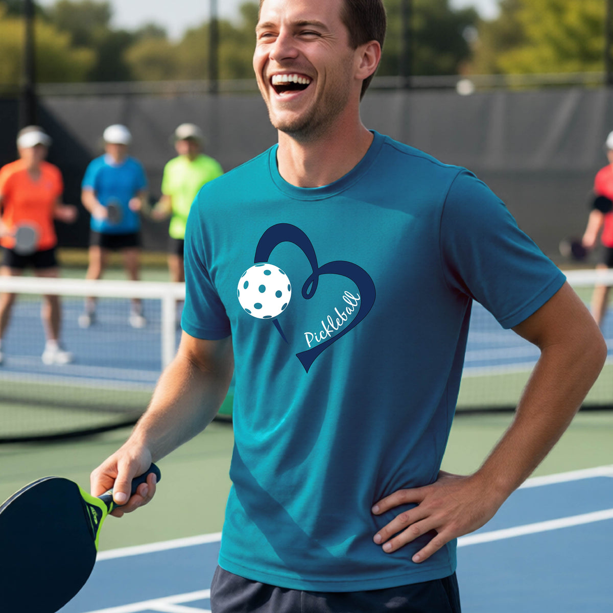 A man laughing on a pickleball court wearing a teal green t-shirt with a navy blue heart, white pickleball graphic, and the text 'pickleball' printed on the front.