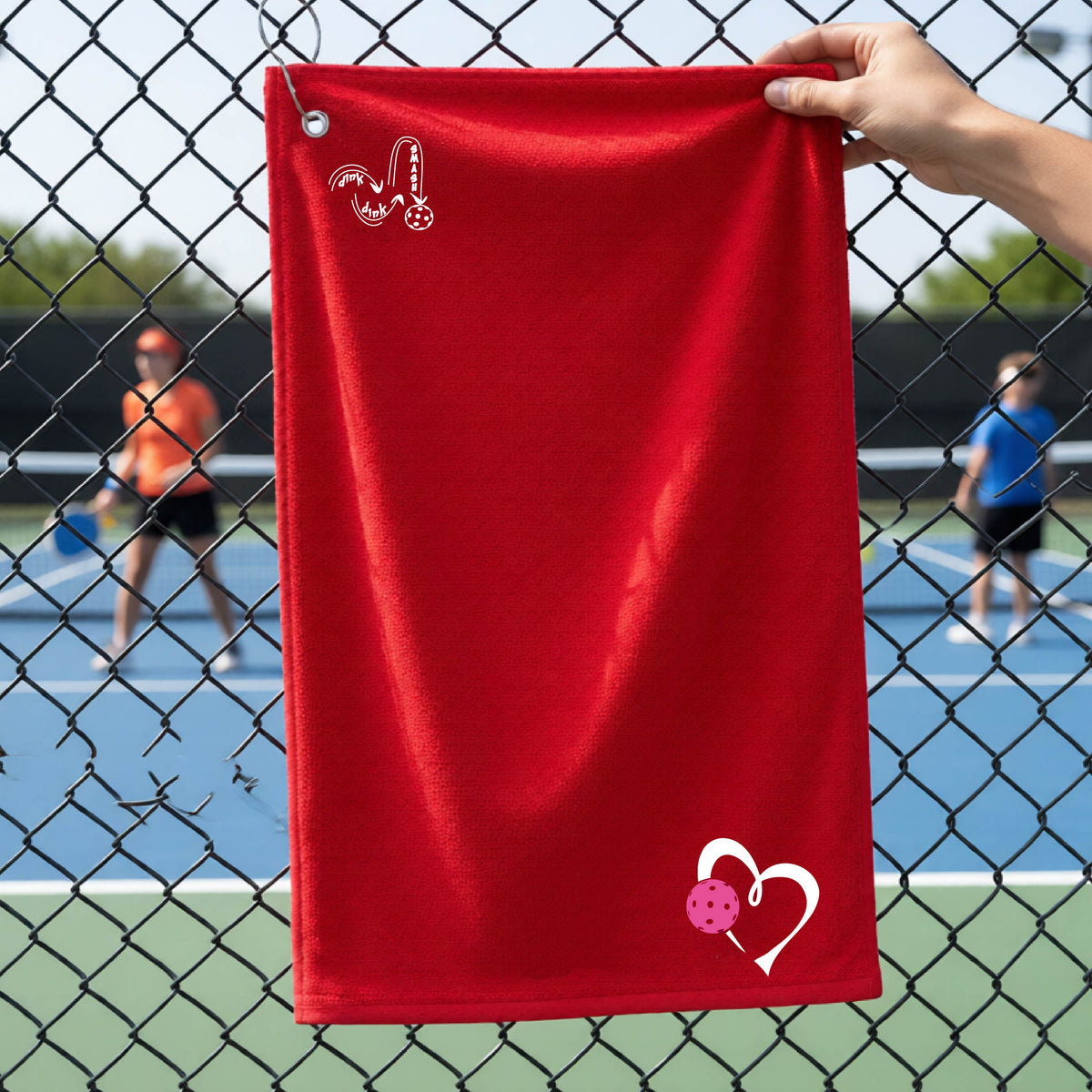 A red textured terry cloth towel held up in front of a chain-link fence at a pickleball court, featuring a white 'DINK DINK SMASH' graphic near the top left and a white heart with a pink pickleball near the bottom right.