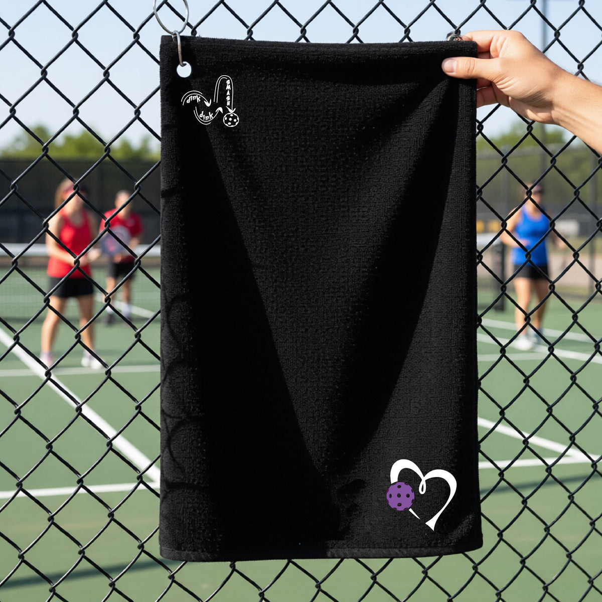 A black athletic terry towel held up by a hand in front of a chain-link fence at a pickleball court. The towel features a white stylized logo 'DINK DINK SMASH' near the top left and a white heart with a purple pickleball ball graphic near the bottom right.
