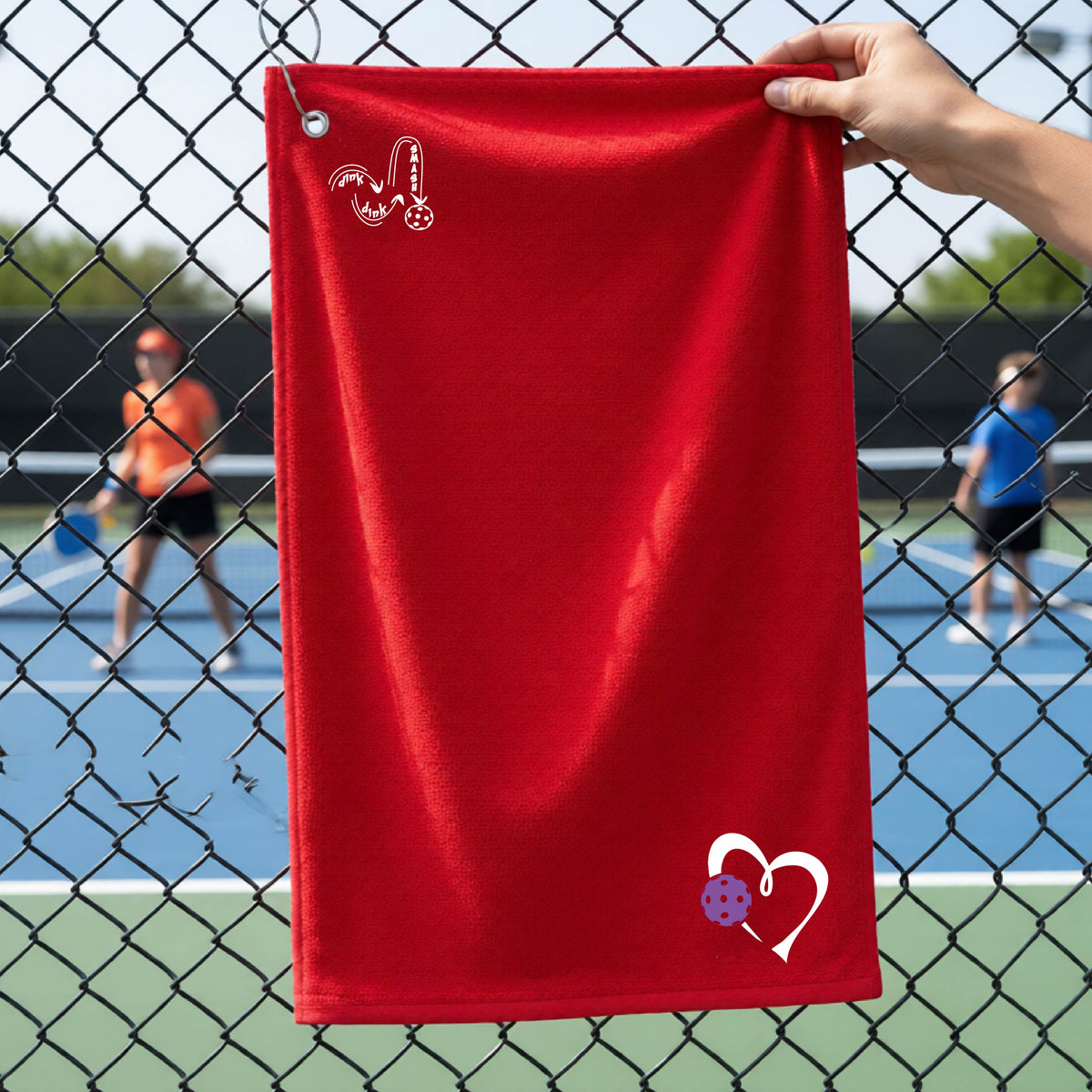 A red athletic terry towel held up by a hand in front of a chain-link fence at a pickleball court. The towel features a white stylized logo 'DINK DINK SMASH' near the top left and a white heart with a purple pickleball ball graphic near the bottom right.