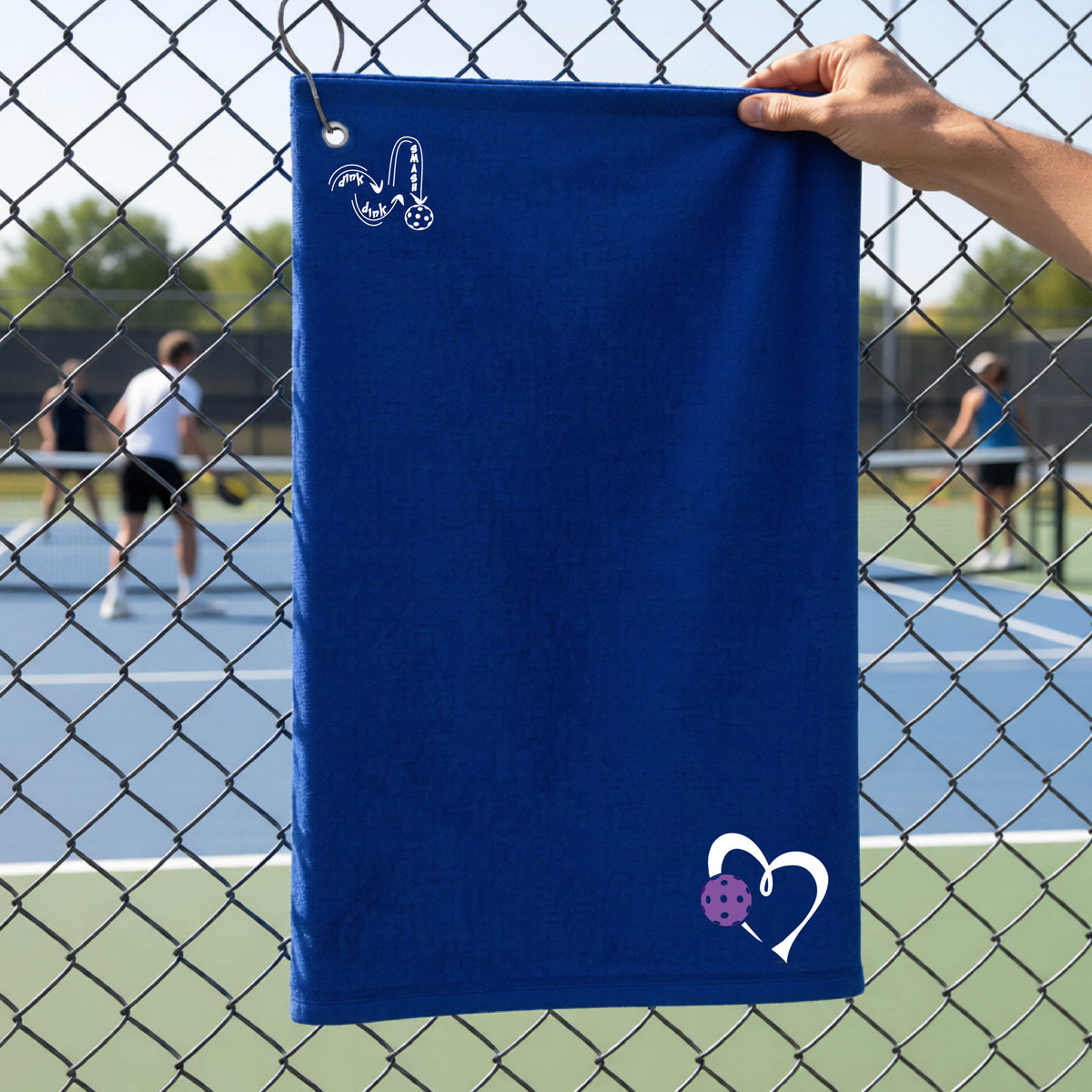 A royal  blue athletic terry towel held up by a hand in front of a chain-link fence at a pickleball court. The towel features a white stylized logo 'DINK DINK SMASH' near the top left and a white heart with a purple pickleball ball graphic near the bottom right.