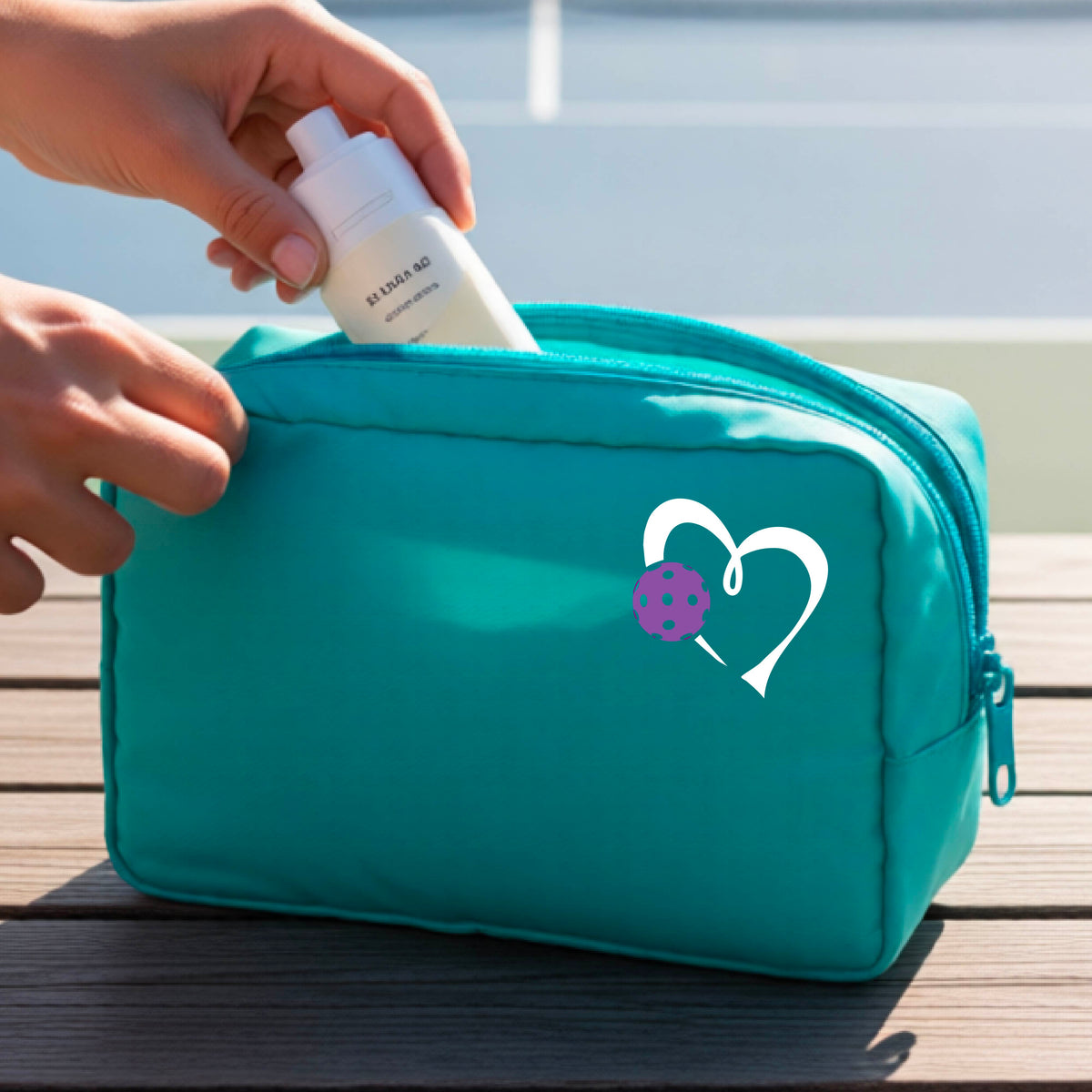 A turquoise zippered toiletry bag sits on a wooden table outdoors, with a person's hands reaching into the open top. A white heart graphic with a purple pickleball accent is printed on the front.