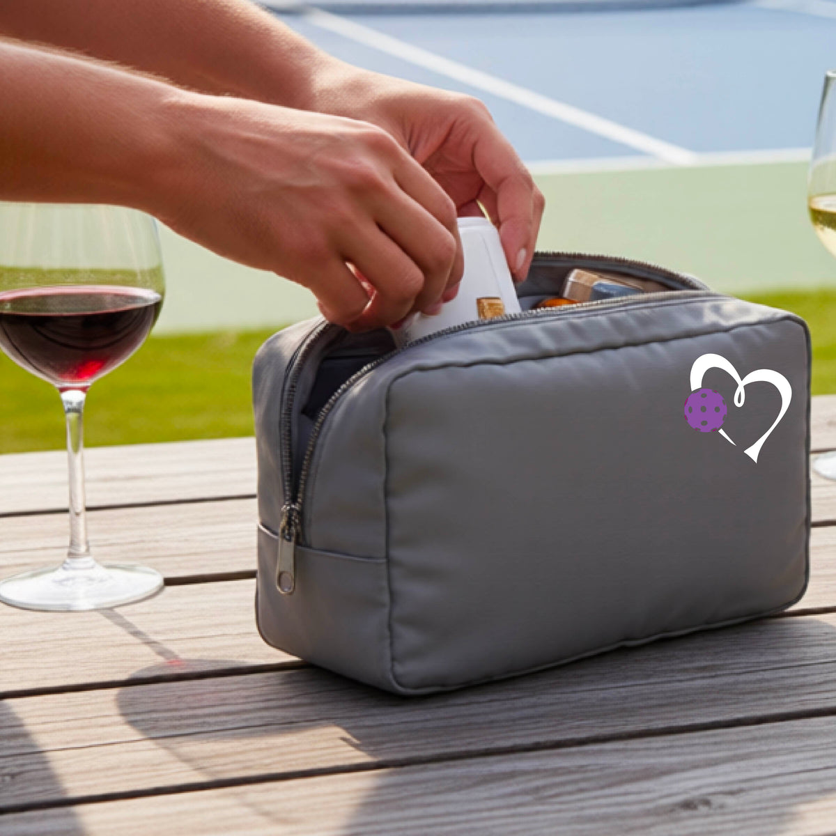 A gray zippered toiletry bag sits on a wooden table outdoors, with a person's hands reaching into the open top. A white heart graphic with a purple pickleball accent is printed on the front.