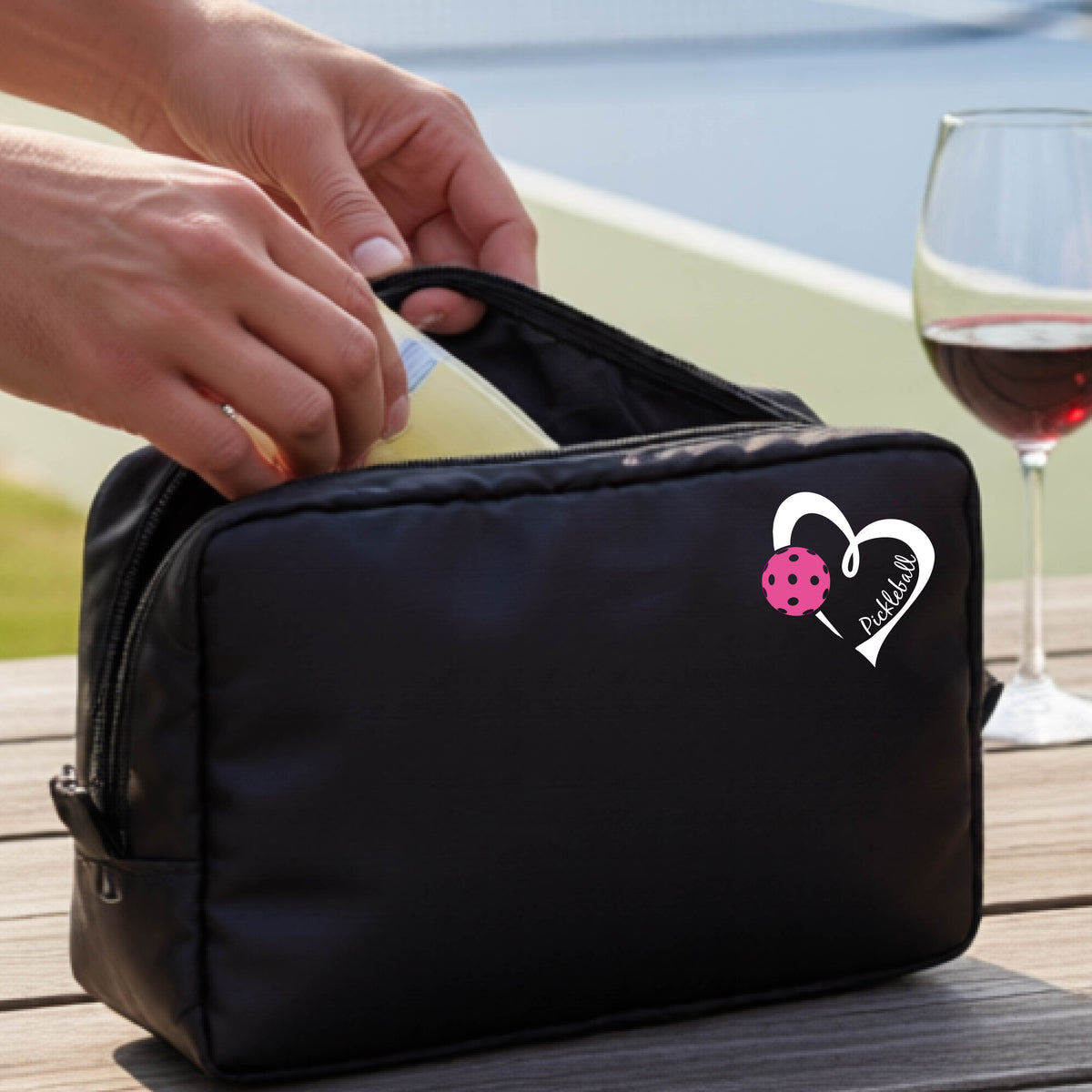 A black zippered cosmetic pouch with a white and pink heart-shaped pickleball  graphic, shown on a wooden table with a wine glass and hands placing items inside.