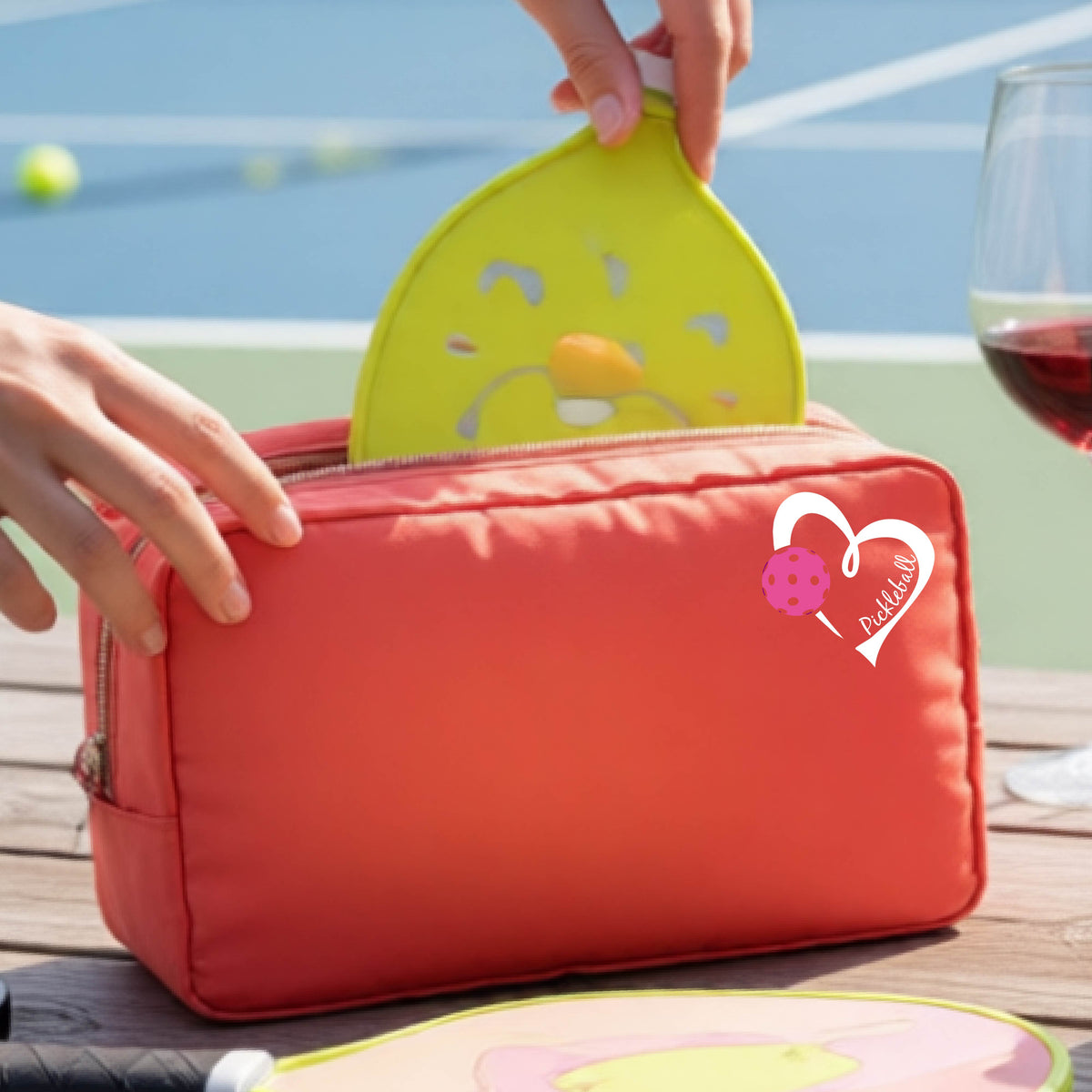 A coral zippered cosmetic pouch with a white and pink heart-shaped pickleball  graphic, shown on a wooden table with a wine glass and hands placing items inside.