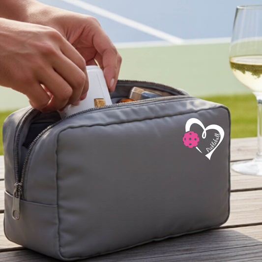 A gray zippered cosmetic pouch with a white and pink heart-shaped pickleball  graphic, shown on a wooden table with a wine glass and hands placing items inside.