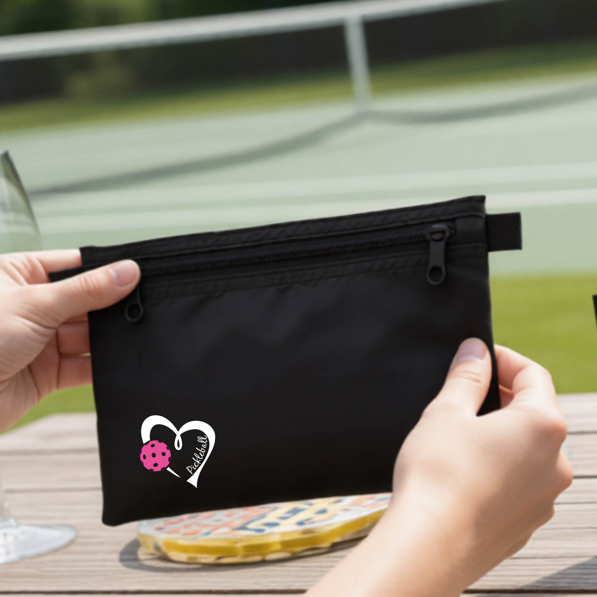 A closeup of a black pickleball small cosmetic zippered bag with a white heart with pink pickleball grpahic and the text 'Pickleball' inside the heart. 