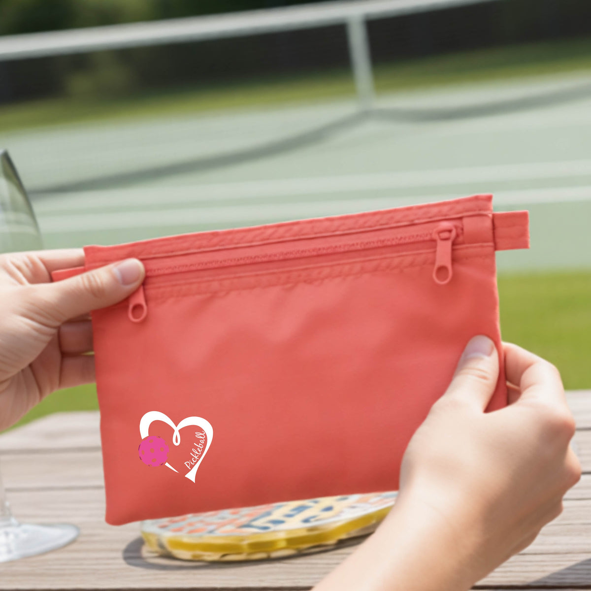 A closeup of a coral pink pickleball small cosmetic zippered bag with a white heart with pink pickleball grpahic and the text 'Pickleball' inside the heart. 