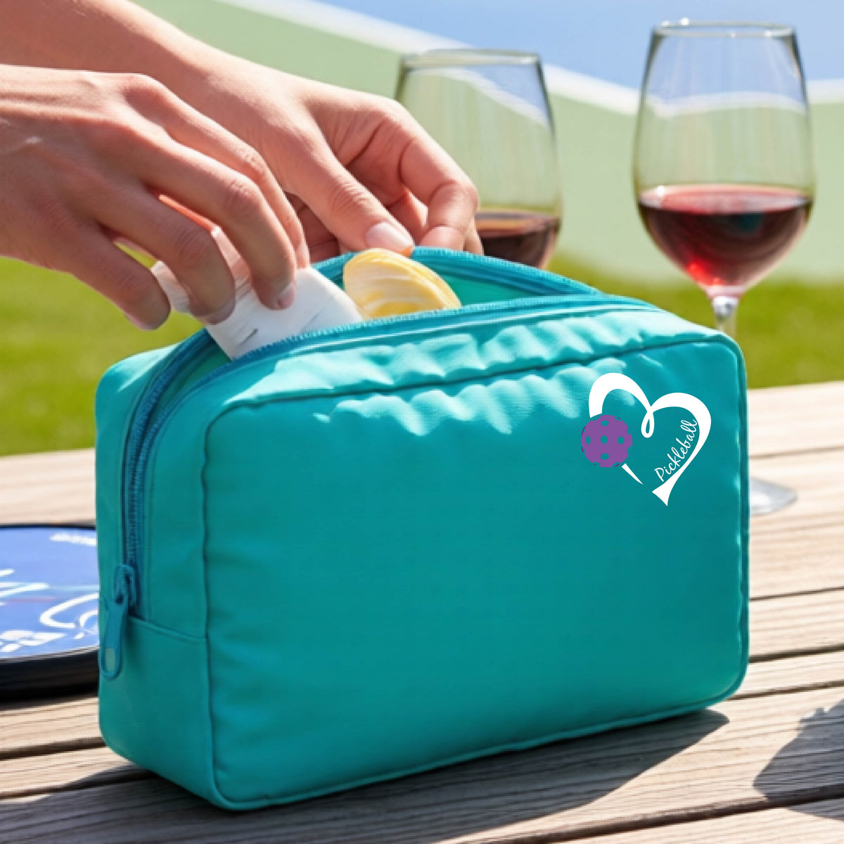 A turquoise zippered cosmetic pouch with a white and purple heart-shaped pickleball  graphic, shown on a wooden table with a wine glass and hands placing items inside.