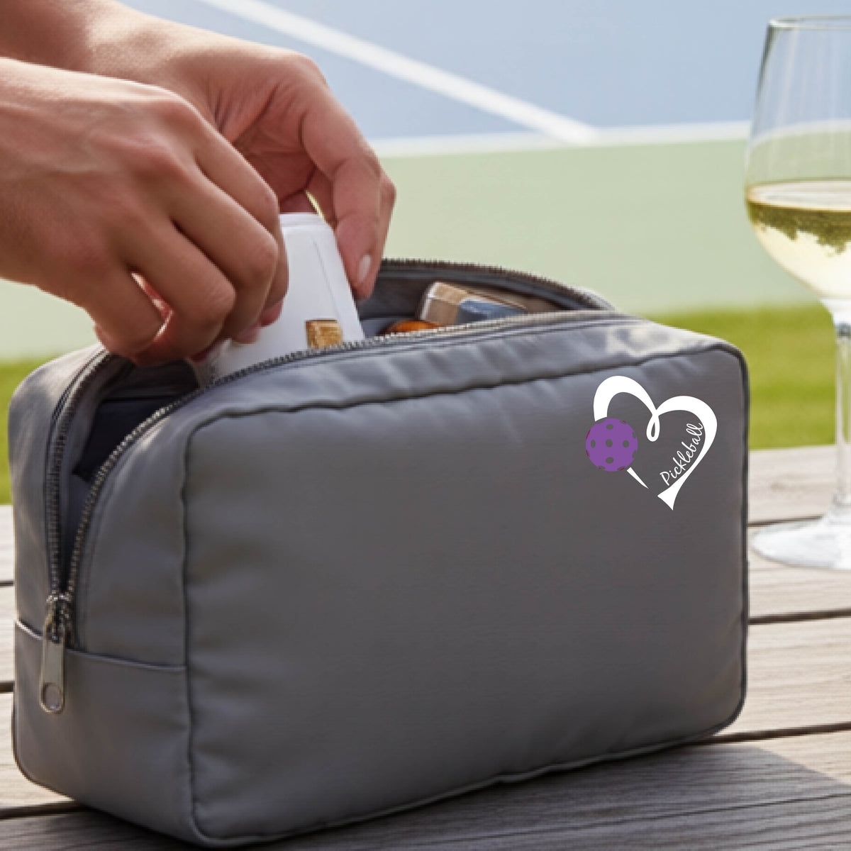 A gray zippered cosmetic pouch with a white and purple heart-shaped pickleball  graphic, shown on a wooden table with a wine glass and hands placing items inside.