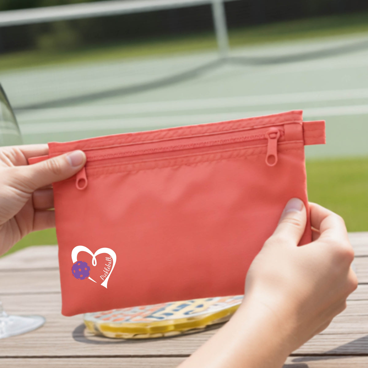 A closeup of a coral pink pickleball small cosmetic zippered bag with a white heart with purple pickleball grpahic and the text 'Pickleball' inside the heart. 