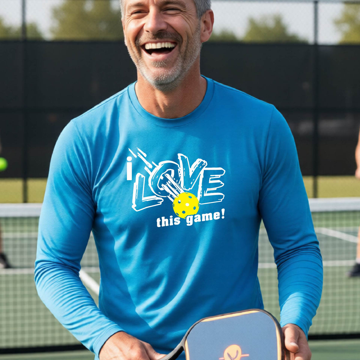 A man wearing a bright blue long-sleeve athletic shirt with a graphic that reads 'i LOVE this game!' in white, with a yellow pickleball motif, standing on a blue pickleball court.