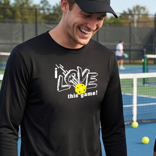 A man wearing a black long-sleeve athletic shirt with a graphic that reads 'i LOVE this game!' in white, with a yellow pickleball motif, standing on a blue pickleball court.