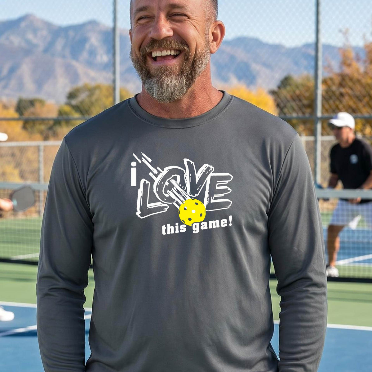 A man wearing a dark gray long-sleeve athletic shirt with a graphic that reads 'i LOVE this game!' in white, with a yellow pickleball motif, standing on a blue pickleball court.
