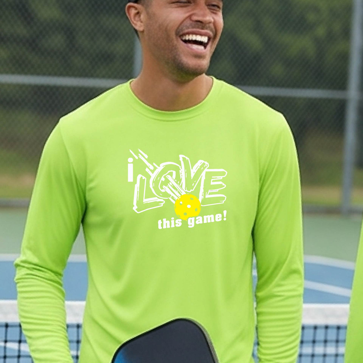 A man wearing a lime-green long-sleeve athletic shirt with a graphic that reads 'i LOVE this game!' in white, with a yellow pickleball motif, standing on a blue pickleball court.