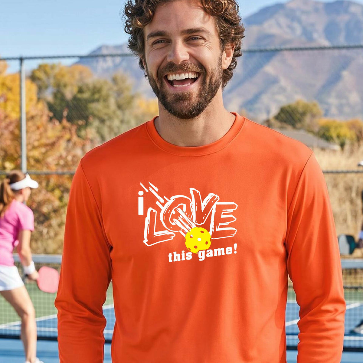 A man wearing a bright orange long-sleeve athletic shirt with a graphic that reads 'i LOVE this game!' in white, with a yellow pickleball motif, standing on a blue pickleball court.