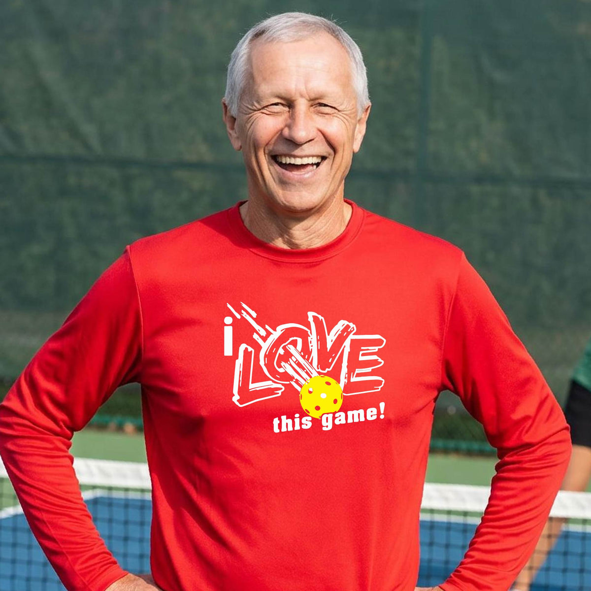 A man wearing a red long-sleeve athletic shirt with a graphic that reads 'i LOVE this game!' in white, with a yellow pickleball motif, standing on a blue pickleball court.