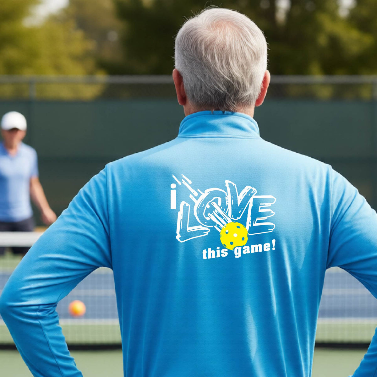 A man on a pickelball court seen from behind wearing a bright blue long-sleeve quarter-zip pullover with a white graphic that reads 'i LOVE this game!' and a yellow oickleball illustration on the back.