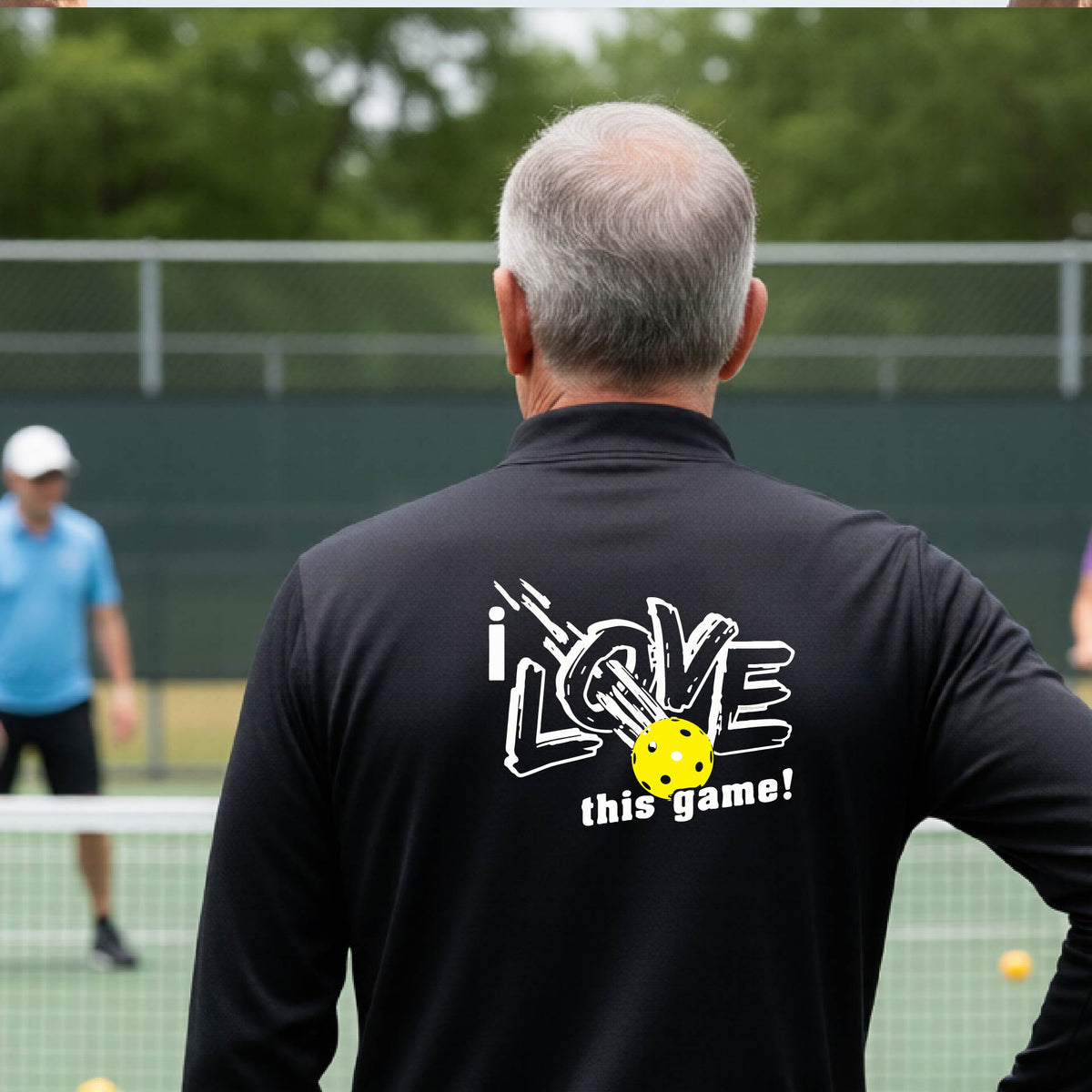 A man on a pickelball court seen from behind wearing a black long-sleeve quarter-zip pullover with a white graphic that reads 'i LOVE this game!' and a yellow oickleball illustration on the back.