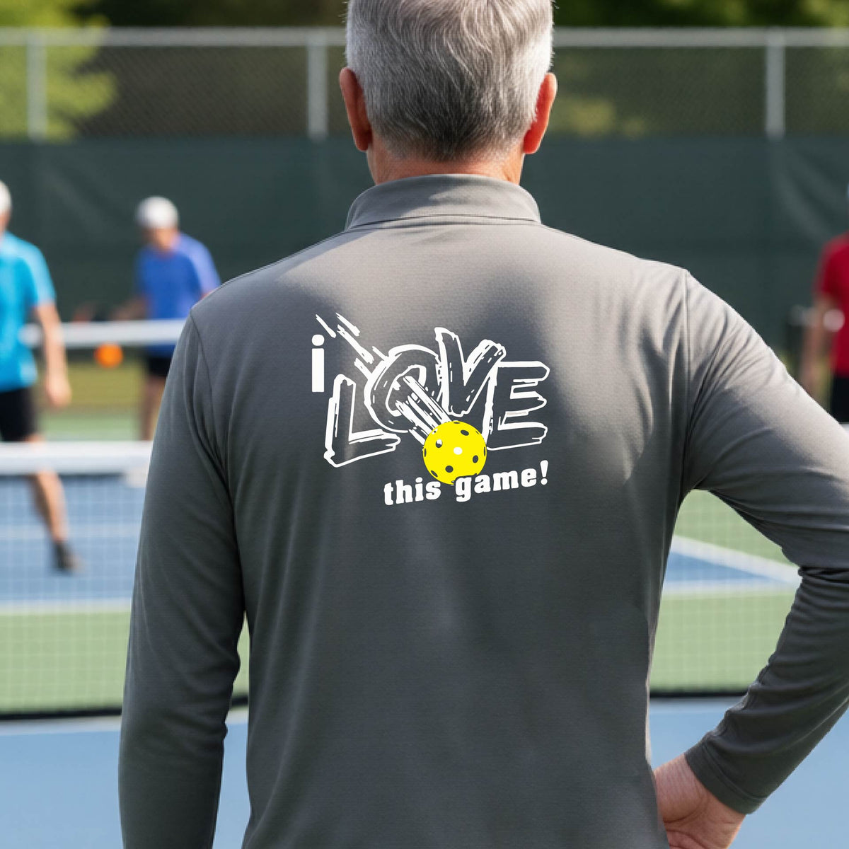 A man on a pickelball court seen from behind wearing a gray long-sleeve quarter-zip pullover with a white graphic that reads 'i LOVE this game!' and a yellow oickleball illustration on the back.