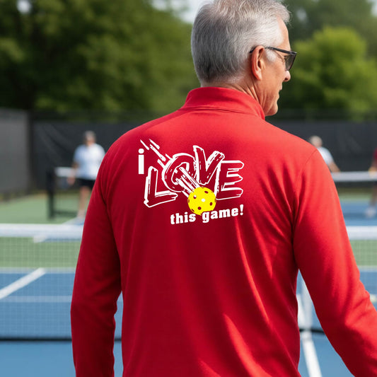 A man on a pickelball court seen from behind wearing a red long-sleeve quarter-zip pullover with a white graphic that reads 'i LOVE this game!' and a yellow oickleball illustration on the back.