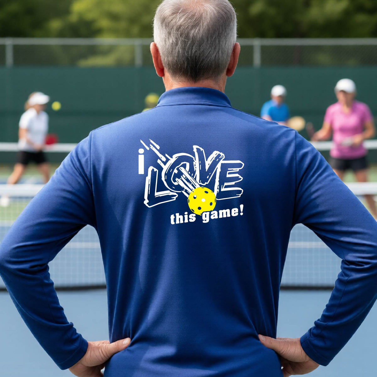 A man on a pickelball court seen from behind wearing a royal blue long-sleeve quarter-zip pullover with a white graphic that reads 'i LOVE this game!' and a yellow oickleball illustration on the back.