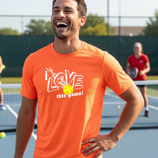A man laughing on an outdoor pickleball court wearing a bright orange t-shirt with a white graphic that reads 'i LOVE this game!' with a yellow pickelball detail.