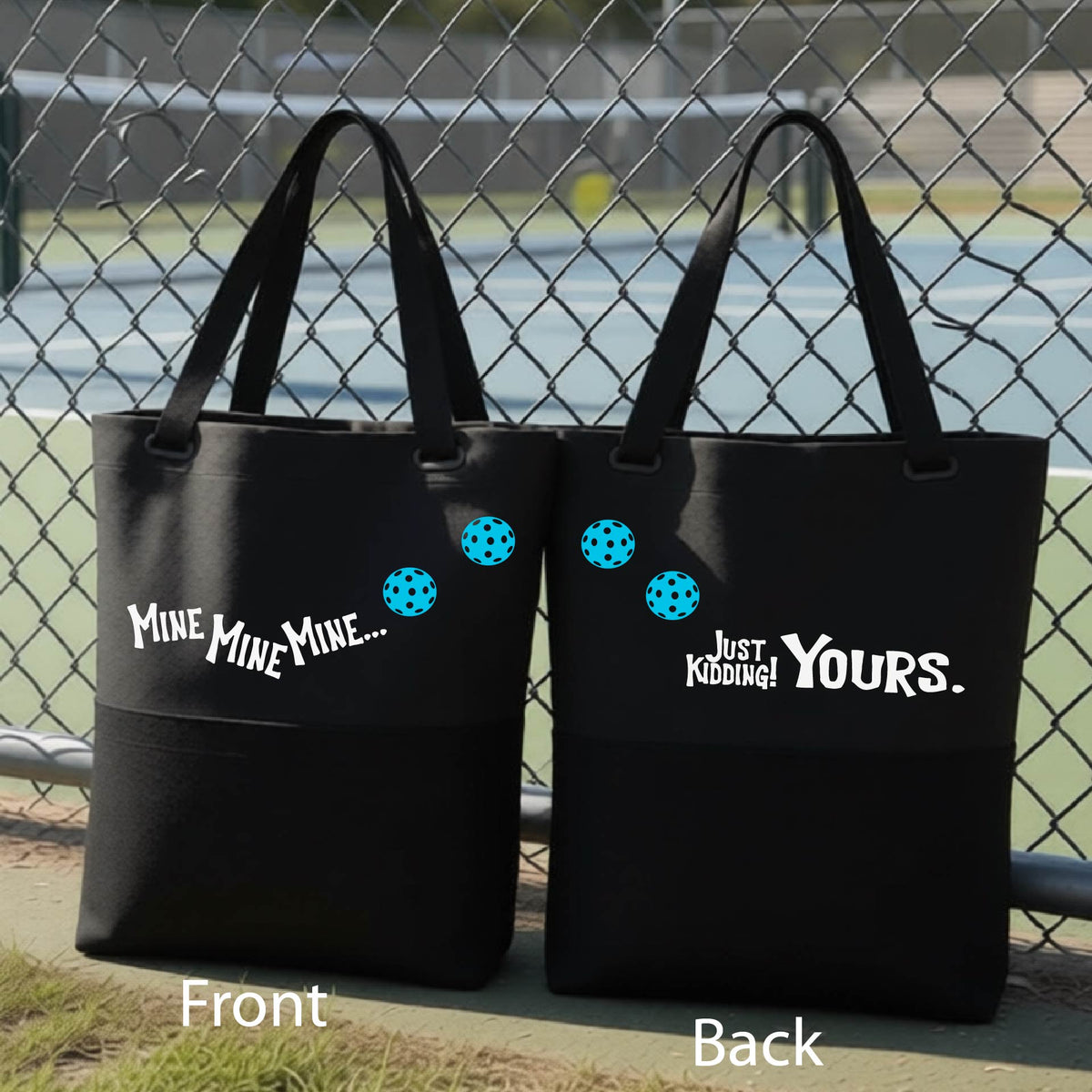A close-up of two black tote bags.  One shows the front with the text 'MINE MINE MINE' along with three pickleball graphics. and the back text 'JUST KIDDING YOURS, along with three pickleball graphics, in front of a pickleball court. 