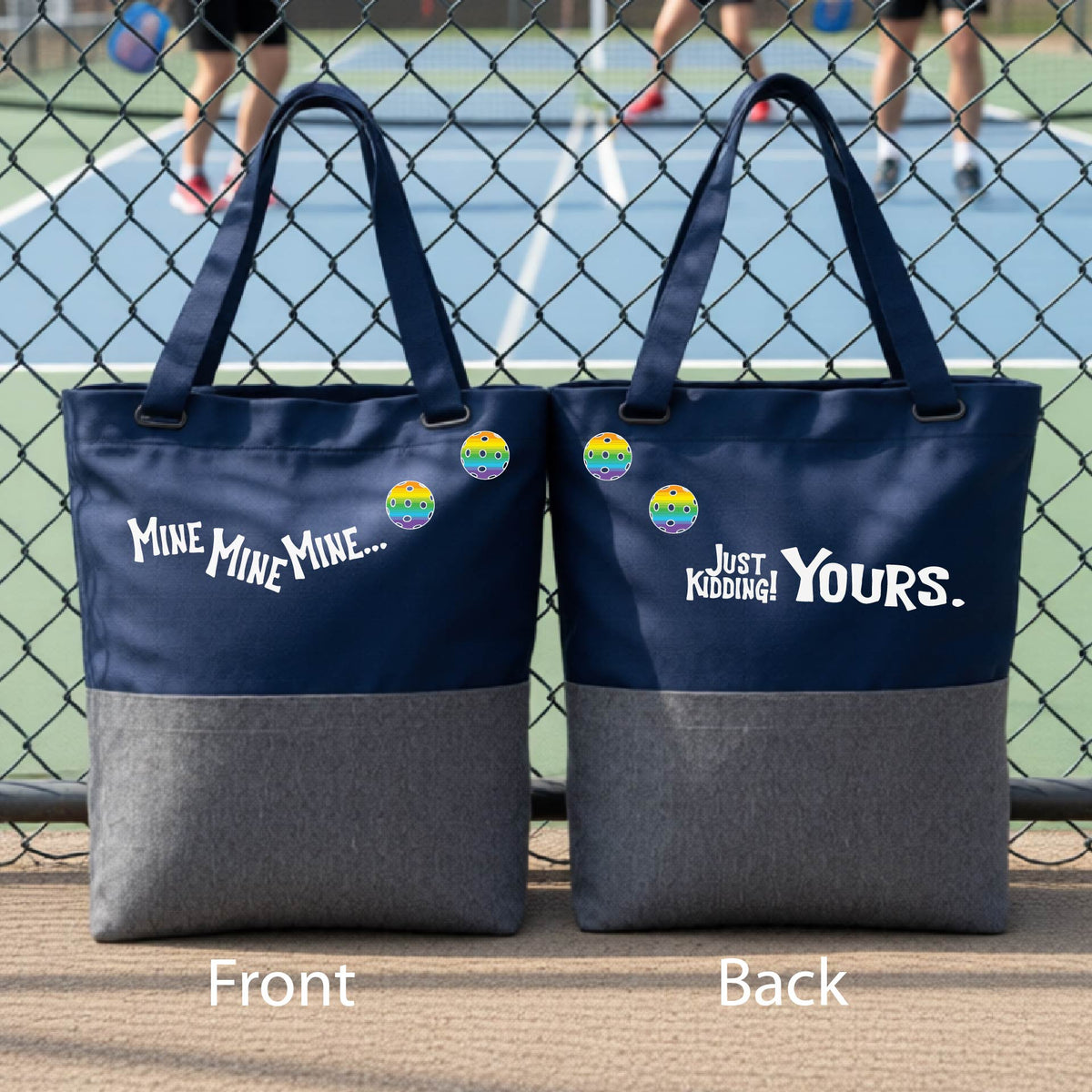 A close-up of two blue and gray tote bags.  One shows the front with the text 'MINE MINE MINE' along with three pickleball graphics. and the back text 'JUST KIDDING YOURS, along with three pickleball graphics, in front of a pickleball court. 