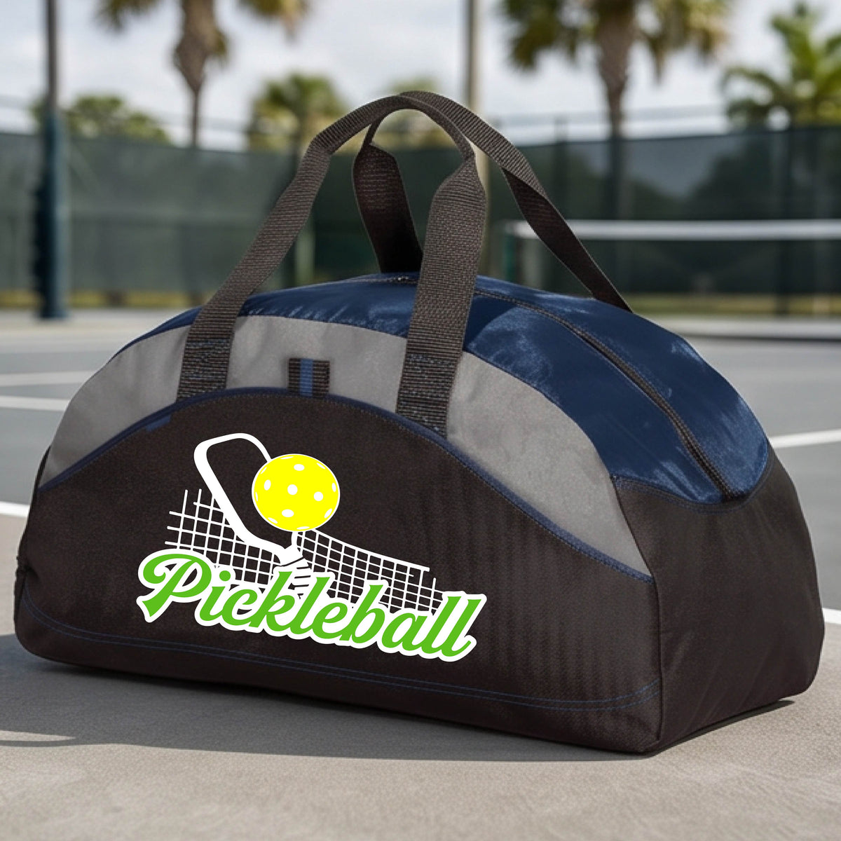 A close-up of a navy blue, black and gray tote bag with a graphic on the front that has white and green text "PICKLEBALL'  with a white pickleball paddle and net and a yellow pickleball above the text graphic.