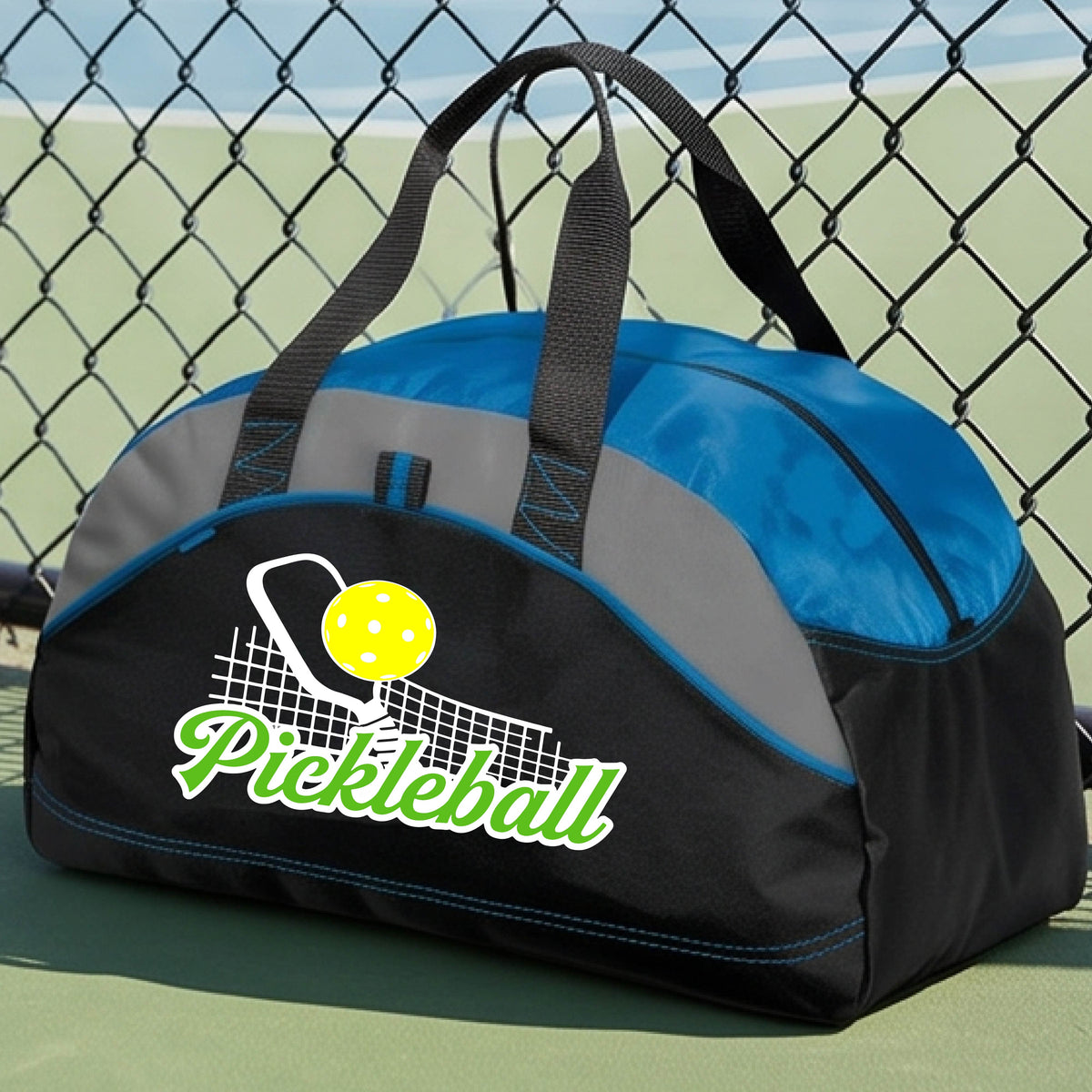 A close-up of a royal blue, black and gray tote bag with a graphic on the front that has white and green text "PICKLEBALL'  with a white pickleball paddle and net and a yellow pickleball above the text graphic.