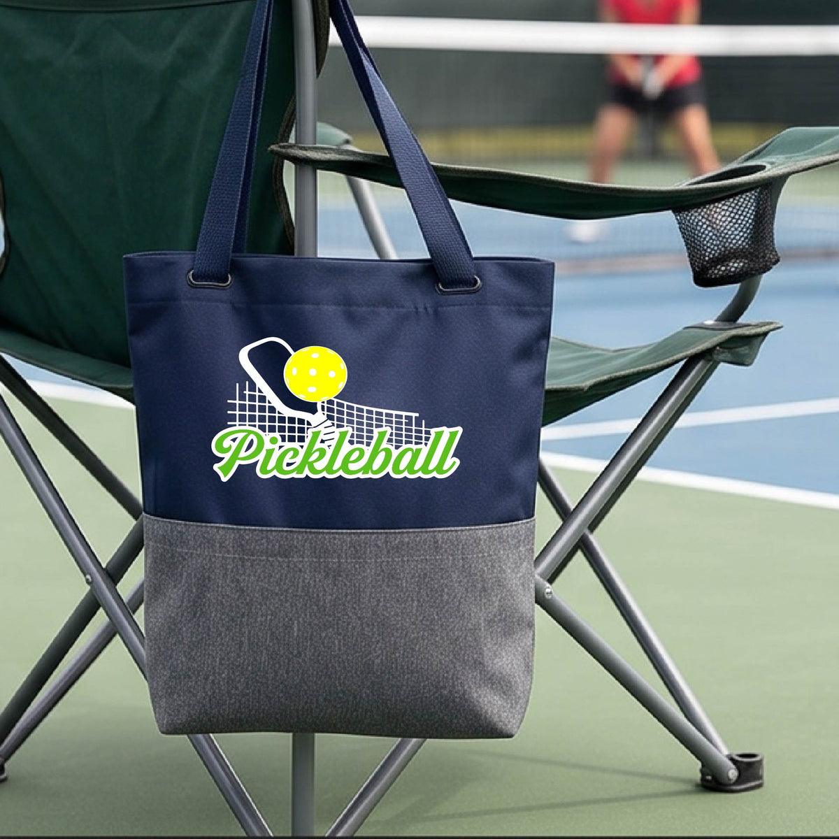 A close-up of a blue and gray tote bag with a graphic on the front that has white and green text "PICKLEBALL'  with a white pickleball paddle and net and a yellow pickleball above the text graphic.