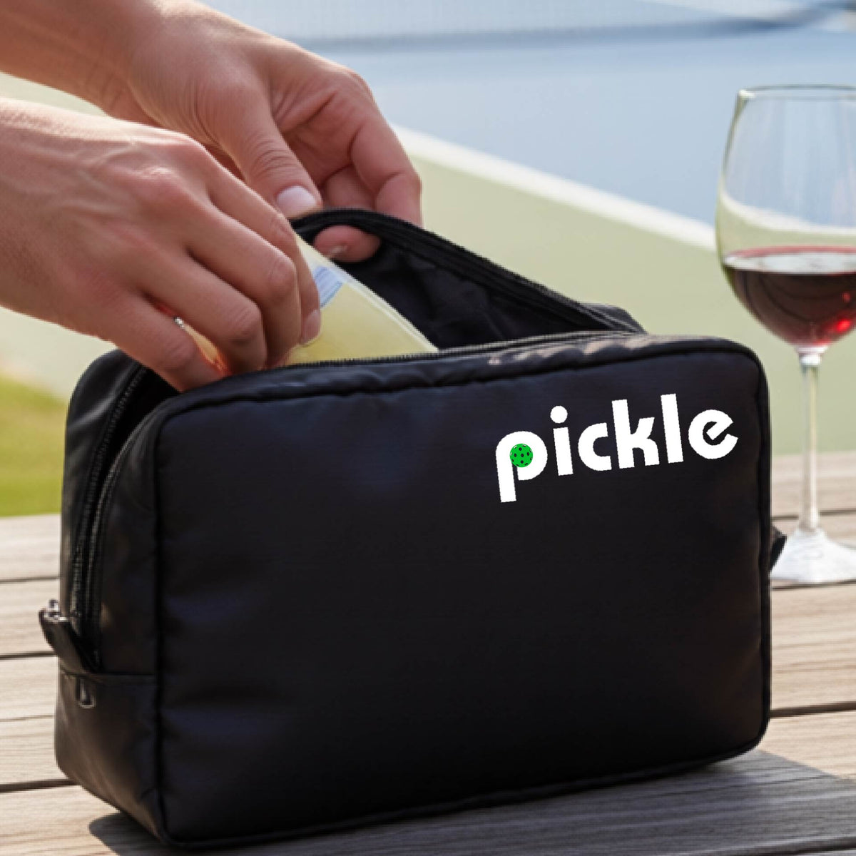 A close-up of a black pickleball polyester cosmetic bag with a white graphic of ‘PICKLE’ with a green pickleball integrated into the letter P printed on the upper right corner, in front of a pickleball court. 