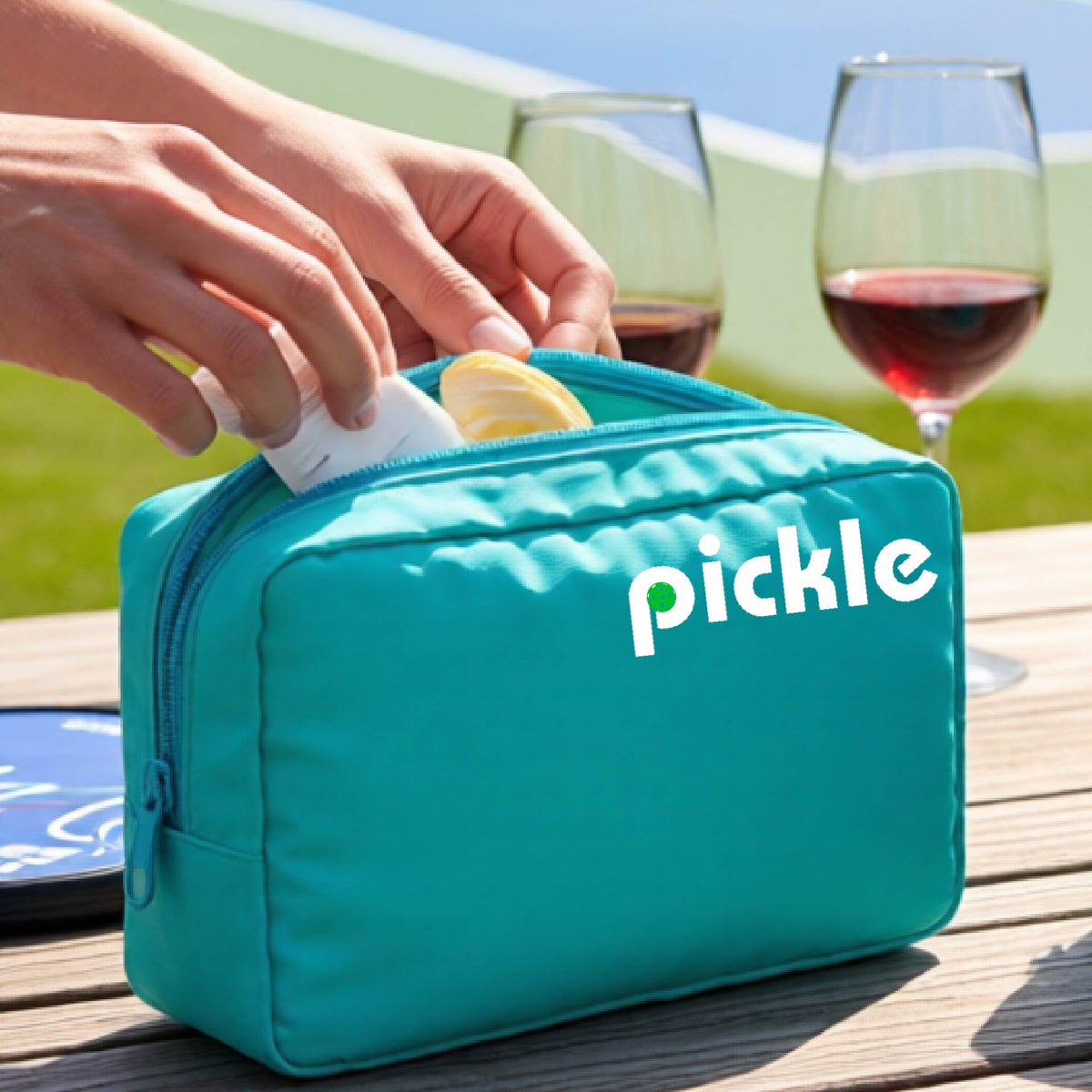 A close-up of a turquoise pickleball polyester cosmetic bag with a white graphic of ‘PICKLE’ with a green pickleball integrated into the letter P printed on the upper right corner, in front of a pickleball court. 