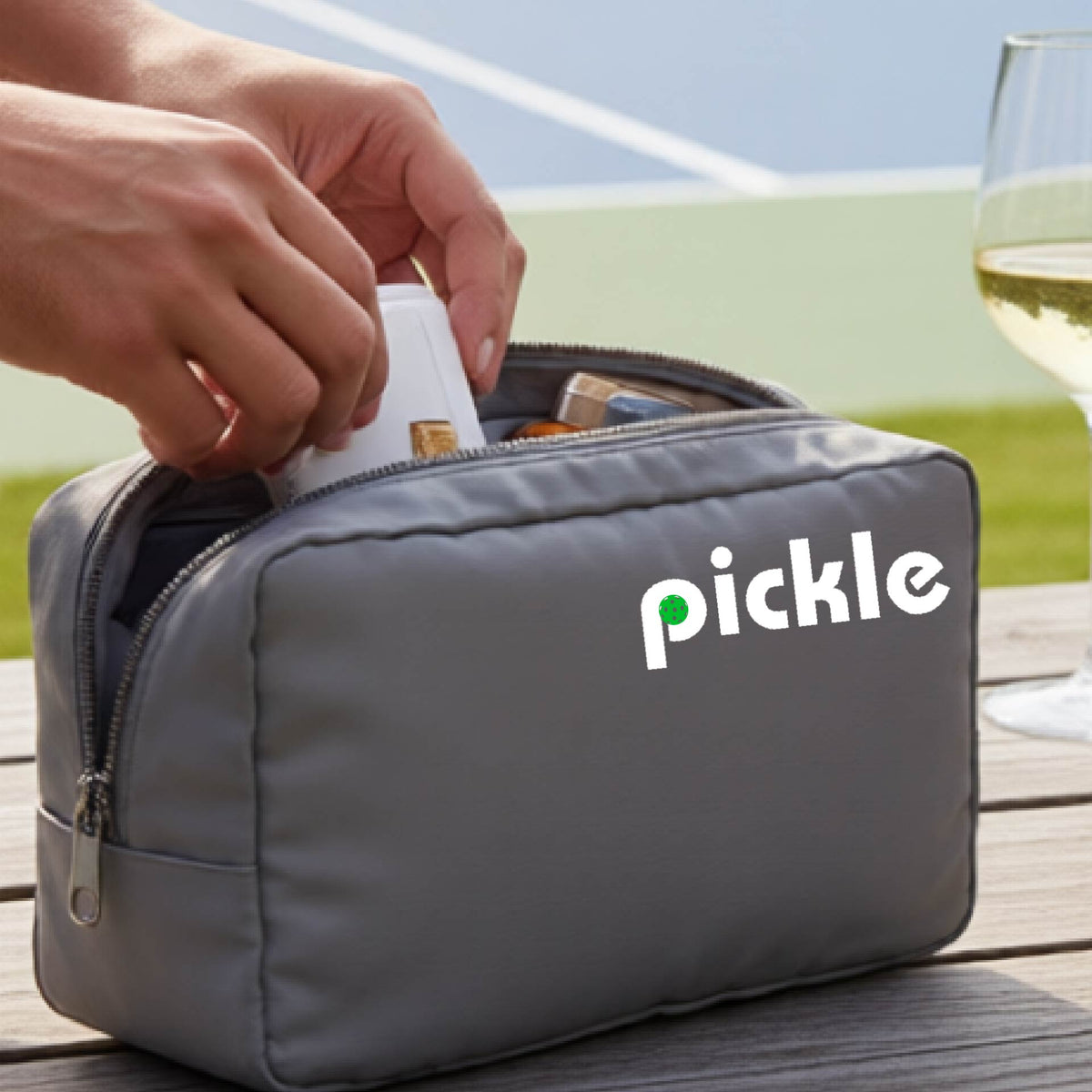 A close-up of a gray pickleball polyester cosmetic bag with a white graphic of ‘PICKLE’ with a green pickleball integrated into the letter P printed on the upper right corner, in front of a pickleball court. 